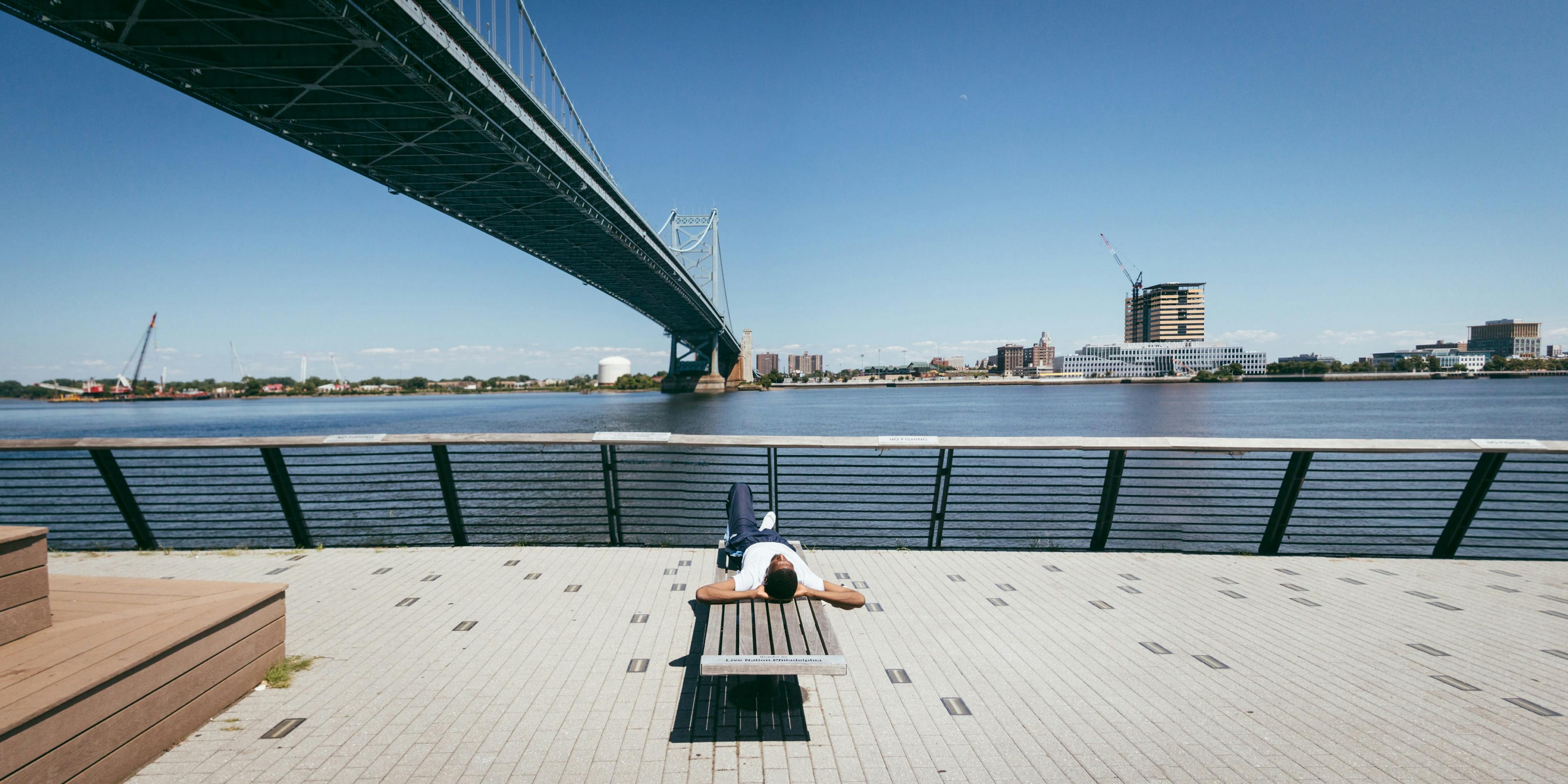 Man laying down on a bench on the Race St Pier in Philadelphia. View looking at the Delaware River.