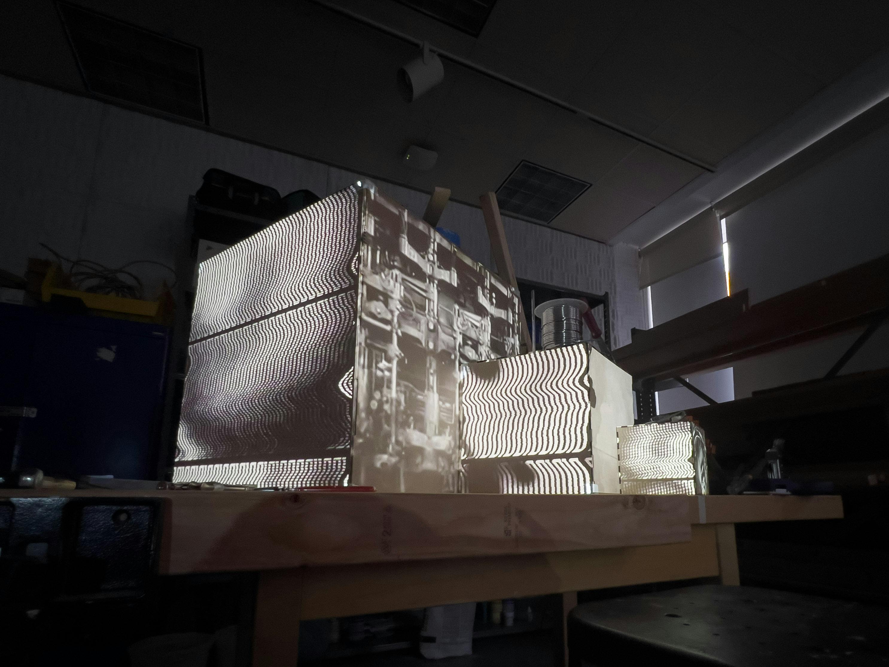Black and white video of a factory projected onto the three cardboard cubes